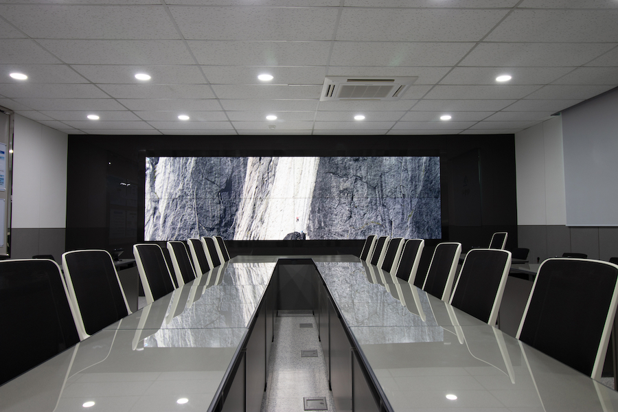 Modern conference room with a large video wall display, sleek glass table, and black executive chairs arranged in rows.