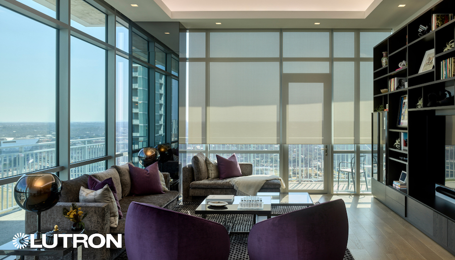 Motorized shades are half-lowered on a window wall in a modern apartment living room overlooking a cityscape.