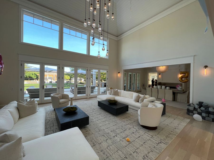 Spacious modern living room with high ceilings, large windows, and glass doors opening to a patio and garden. The room features white sofas, a light area rug, black coffee tables, and hanging pendant lights.