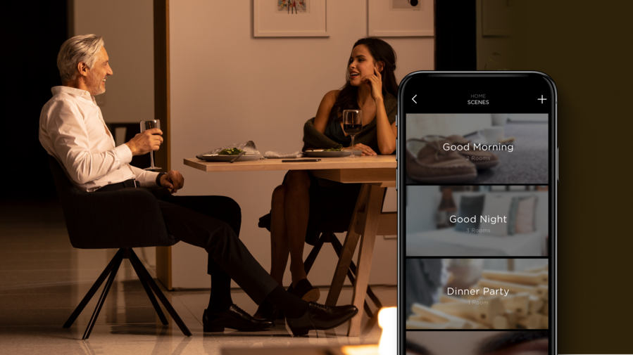 A couple sits at a dining table in a dimly lit room. A Savant app interface is in the foreground.