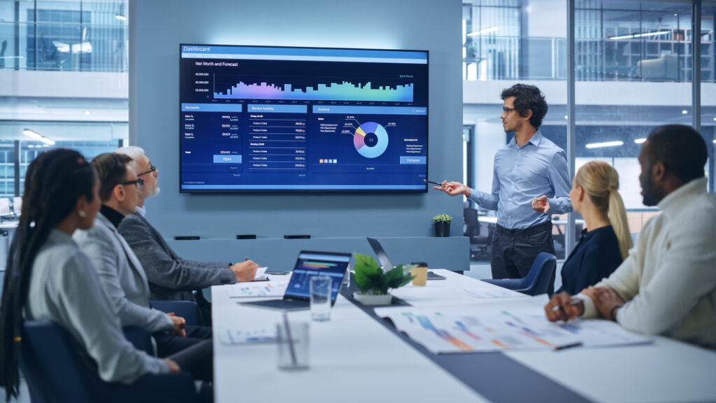 Business team in a modern conference room watching a presenter share data on a large screen during a meeting.