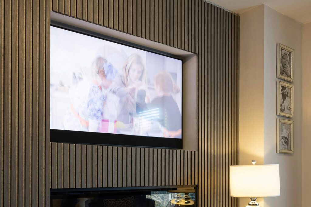 Wall-mounted TV recessed in slatted wood paneling above fireplace in cozy living space