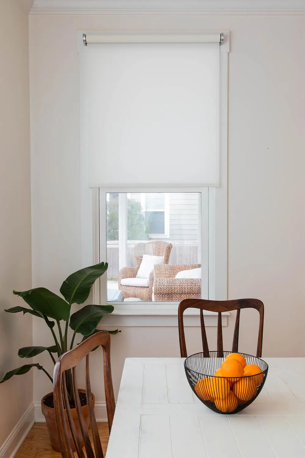 Comedor minimalista con mesa blanca, frutero con naranjas, dos sillas de madera y vista a un área exterior con muebles de mimbre.