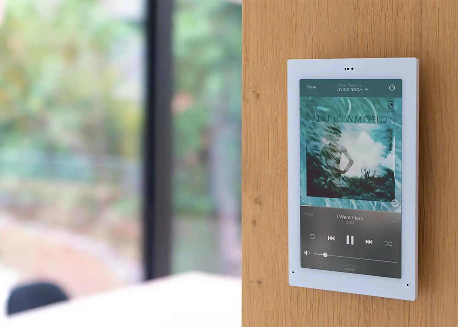 That wall‑mounted touchscreen is showing a music player interface with album art, track info, and playback controls — a clean smart‑home style display set into a wooden surface with a soft, blurred background of greenery outside.